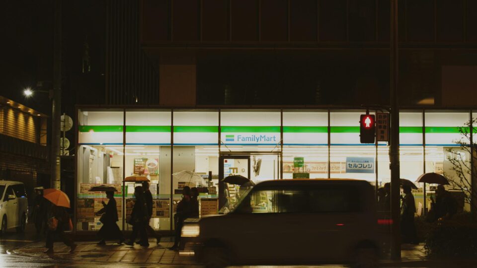 People crossing a street near a FamilyMart in Japan during a rainy night.